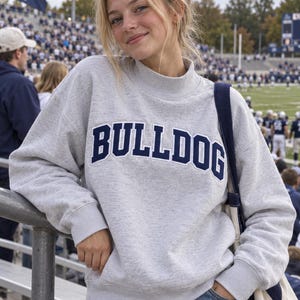 May include: A light grey long-sleeve sweatshirt featuring a mock neck collar and ribbed cuffs. The word BULLDOG is printed across the chest in bold, dark navy blue capital letters with a white outline, creating a collegiate-inspired aesthetic.