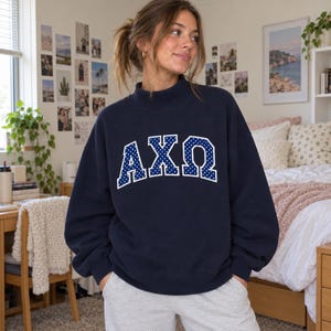 May include: A navy blue long-sleeve sweatshirt with a mock neck collar. The front features the Greek letters AXΩ in a bold, arched font. The letters are navy blue with a white polka dot pattern and a thick white outline.