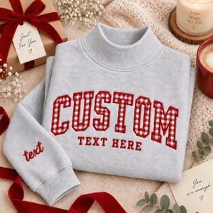 May include: A light grey mock neck sweatshirt featuring the embroidered red and white gingham word CUSTOM in a collegiate font, with the words TEXT HERE embroidered below in red, and the word text embroidered in red on the left sleeve cuff.