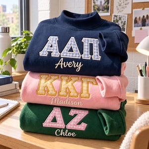 Peut inclure: Une pile de trois sweat-shirts à col montant bleu marine, rose clair et vert foncé. Chacun présente des lettres grecques brodées avec un motif vichy et un prénom personnalisé en écriture cursive : Avery, Madison et Chloe respectivement.
