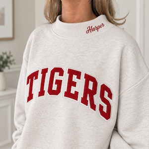 May include: A heathered beige sweatshirt featuring the word TIGERS in large, bold red block letters with a white outline across the chest. The collar area includes the name Harper embroidered in a red cursive script font on the right side.