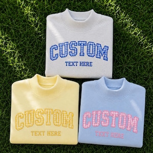 May include: Three mock neck sweatshirts in light gray, pale yellow, and light blue. Each features embroidered text with the word CUSTOM in a gingham pattern and the words TEXT HERE underneath in a matching solid color thread.