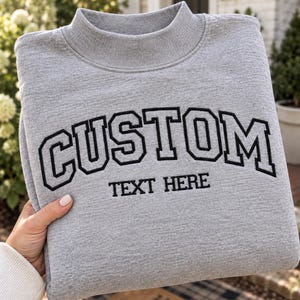 May include: A heather grey mock neck sweatshirt featuring the word CUSTOM embroidered in large black collegiate-style lettering across the chest, with the words TEXT HERE embroidered in a smaller black font directly underneath the main text.