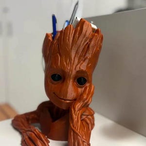 Può includere: Una statuetta di Groot stampata in 3D di colore marrone con un viso sorridente, che tiene una penna in mano. La statuetta è un organizzatore da scrivania con una testa cava per contenere penne e matite.