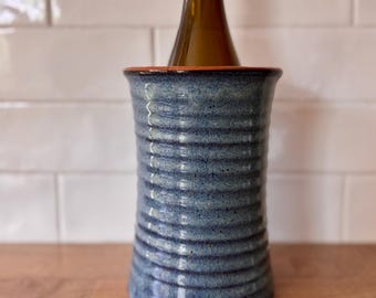 May include: A blue ceramic wine cooler with a textured, ribbed design. A wine bottle with a gold neck is inside the cooler. The wine bottle is partially visible, with the label facing forward. The cooler is on a wooden surface.