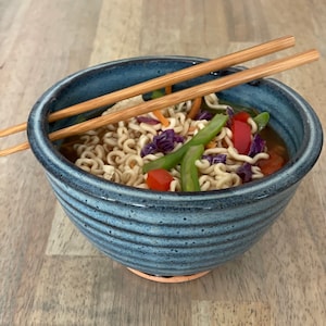 May include: A blue ceramic bowl filled with ramen noodles, vegetables, and broth. The bowl has a textured surface and is resting on a wooden table. Two wooden chopsticks are placed across the top of the bowl. The vegetables include red bell peppers, green beans, and purple cabbage.