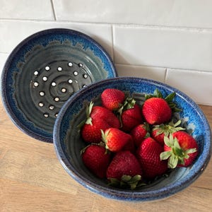 May include: A ceramic colander and bowl set in shades of blue and gray. The colander has a series of white dots. The bowl is filled with fresh, ripe strawberries. The set is on a wooden surface.