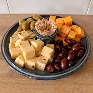 May include: A blue ceramic serving platter with a variety of appetizers. The platter is divided into sections holding green olives, cubed pepper jack cheese, cheddar cheese cubes, and dark red olives. A small bowl of toothpicks sits in the center.