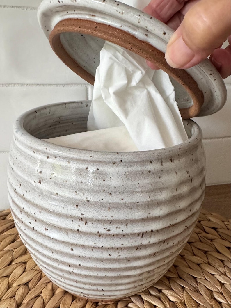 May include: A round, speckled off-white ceramic canister with horizontal ridges and a matching lid. The canister contains white tissues, with one tissue partially pulled through the opening in the lid. The ceramic features a visible brown clay rim.