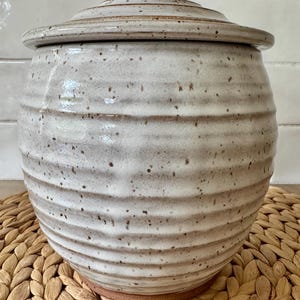 May include: A round ceramic canister with a lid, featuring a cream-colored glaze with brown speckles and horizontal ribbed textures. The base of the canister is exposed brown clay, and the overall shape is bulbous with a slightly tapered neck.