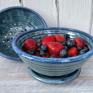 May include: A blue ceramic bowl filled with fresh strawberries and blueberries. A second bowl with drainage holes sits behind it. The bowls are on a matching blue saucer. The fruit is red and blue.