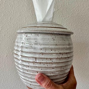 May include: A round, off-white ceramic tissue holder with a speckled finish and horizontal ribbed texture. The container features a matching lid with a central opening, through which a single white paper tissue is pulled upward.