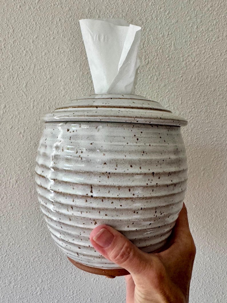 May include: A round, off-white ceramic tissue holder with a speckled finish and horizontal ribbed texture. The container features a matching lid with a central opening, through which a single white paper tissue is pulled upward.