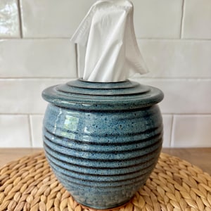 May include: A round, blue ceramic tissue holder with a speckled glaze and horizontal ribbed texture. The container features a matching lid with a central opening, through which a single white tissue is pulled upward, creating a functional display.