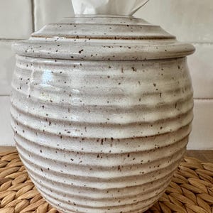 May include: A round, ceramic tissue holder with a glossy, off-white glaze and a speckled brown finish. The container features horizontal, ribbed ridges along its body and a matching lid with a central opening for dispensing white tissues.