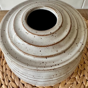 May include: A round, off-white ceramic vase with a speckled finish and a series of horizontal, concentric ridges that taper toward a central circular opening. The glaze features brown flecks throughout, and the rim of the opening is defined by a smooth, dark brown edge.