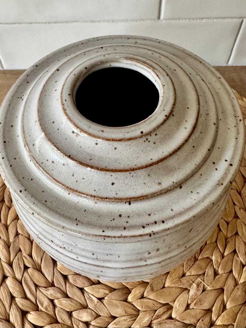 May include: A round, off-white ceramic vase with a speckled finish and a series of horizontal, concentric ridges that taper toward a central circular opening. The glaze features brown flecks throughout, and the rim of the opening is defined by a smooth, dark brown edge.
