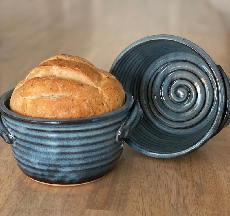 Puede incluir: Una campana de pan de cer&aacute;mica azul con tapa. La campana tiene un dise&ntilde;o de espiral y est&aacute; llena de un pan reci&eacute;n horneado.