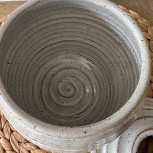 May include: A light gray ceramic bowl with a speckled finish and a visible spiral pattern etched into the interior base. The bowl features a smooth, glossy glaze and a rounded rim, accompanied by a matching circular lid resting to the side.