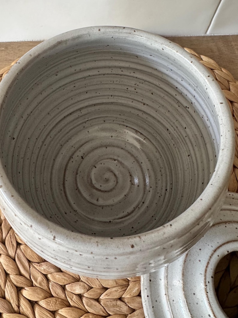 May include: A light gray ceramic bowl with a speckled finish and a visible spiral pattern etched into the interior base. The bowl features a smooth, glossy glaze and a rounded rim, accompanied by a matching circular lid resting to the side.