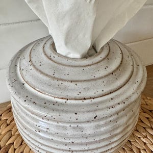 May include: A round, ceramic tissue box cover featuring a light gray, speckled glaze with a series of horizontal, concentric ridges circling the exterior. A single white tissue is pulled through the circular opening at the top of the container.