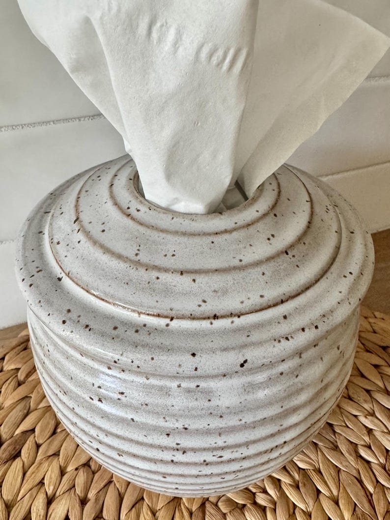 May include: A round, ceramic tissue box cover featuring a light gray, speckled glaze with a series of horizontal, concentric ridges circling the exterior. A single white tissue is pulled through the circular opening at the top of the container.