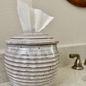 May include: A round, ceramic tissue holder with a white, speckled glaze and horizontal ribbed texture. The container features a matching lid with a central opening, through which a single white facial tissue is pulled upward and out.
