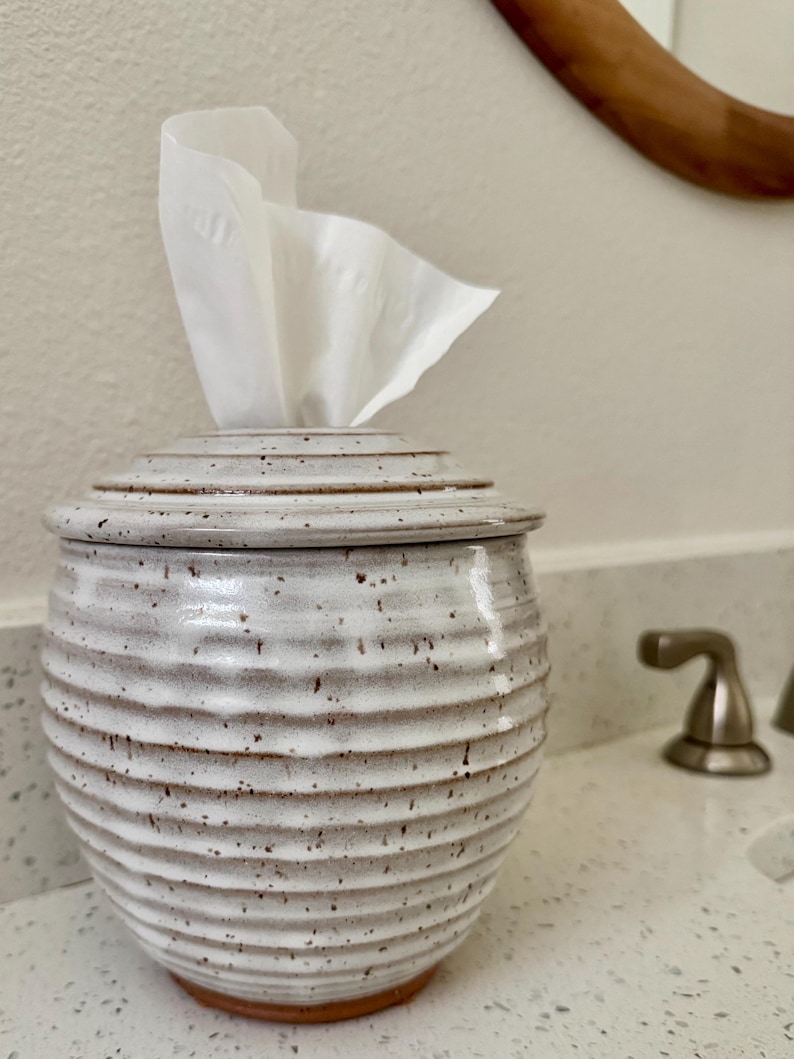 May include: A round, ceramic tissue holder with a white, speckled glaze and horizontal ribbed texture. The container features a matching lid with a central opening, through which a single white facial tissue is pulled upward and out.
