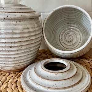 May include: Two off-white ceramic containers with a speckled finish and horizontal ribbed texture. One container holds a white tissue, while the other sits open next to its matching circular lid, which features a central opening and concentric rings.
