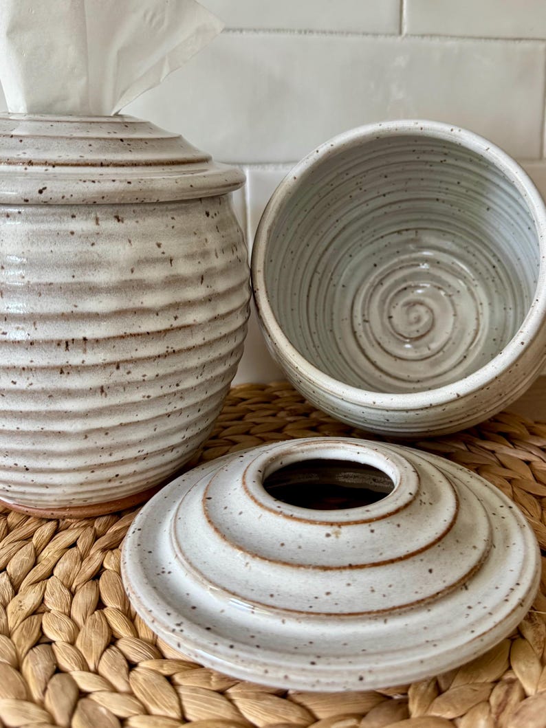 May include: Two off-white ceramic containers with a speckled finish and horizontal ribbed texture. One container holds a white tissue, while the other sits open next to its matching circular lid, which features a central opening and concentric rings.