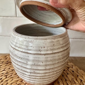 May include: A round, speckled off-white ceramic jar with horizontal ridges and a matching lid. The interior of the lid and the base of the jar reveal the natural, unglazed reddish-brown clay. The jar has a wide opening and a rounded body shape.