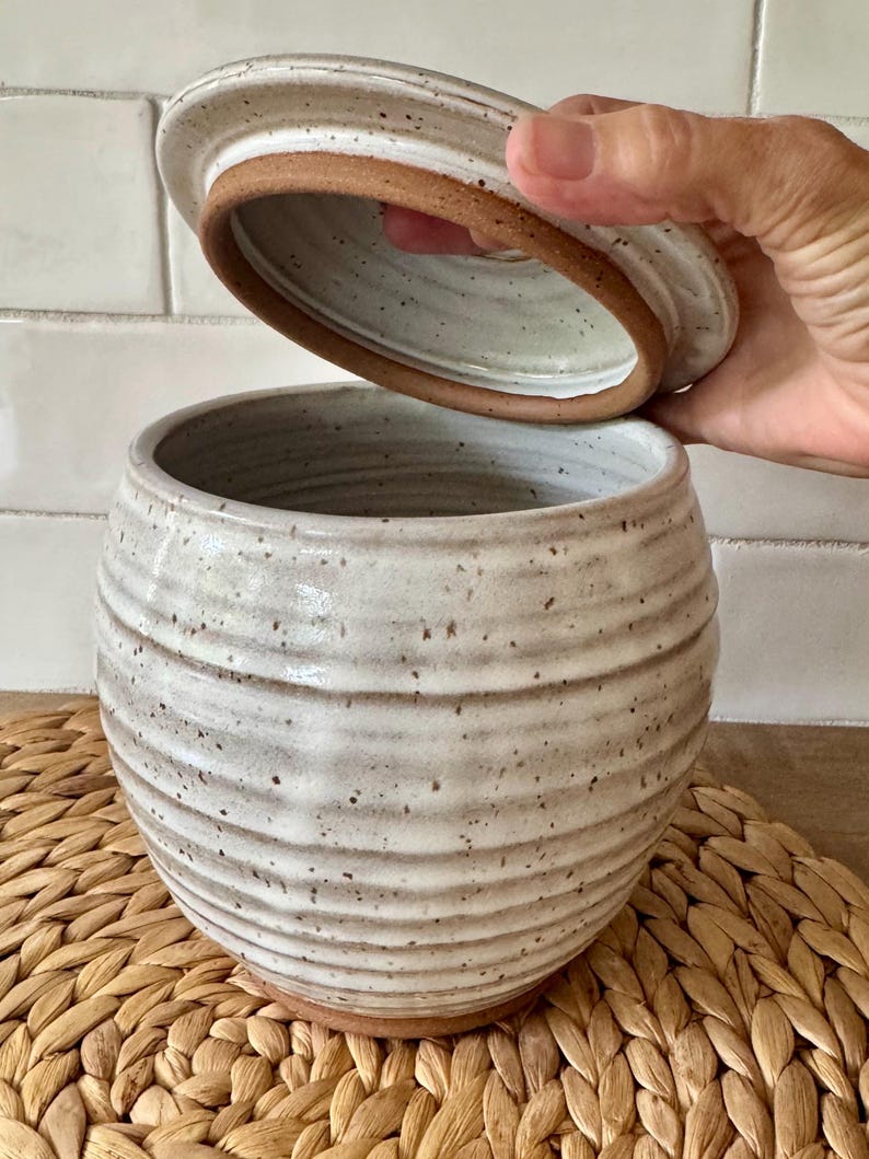 May include: A round, speckled off-white ceramic jar with horizontal ridges and a matching lid. The interior of the lid and the base of the jar reveal the natural, unglazed reddish-brown clay. The jar has a wide opening and a rounded body shape.