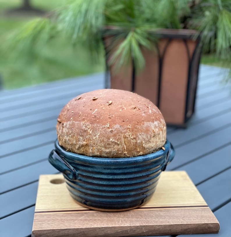 Puede incluir: Una hogaza de pan reci&eacute;n horneada en un bol de cer&aacute;mica azul y blanco. El bol tiene un aspecto r&uacute;stico y artesanal y est&aacute; sobre una tabla de cortar de madera.