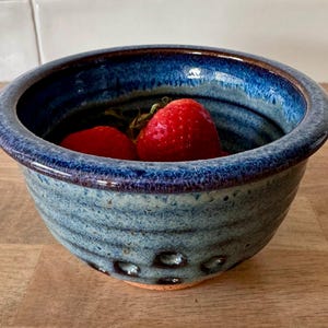 May include: A blue ceramic bowl with a speckled glaze and a rim of dark blue. The bowl is filled with three red strawberries.