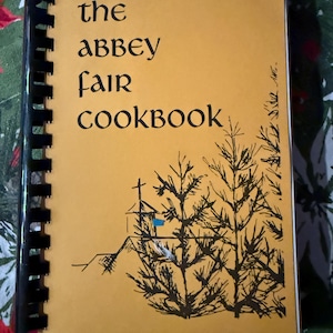 May include: A spiral-bound cookbook titled "The Abbey Fair Cookbook" with a mustard-yellow cover. The cover features black text and a black and white illustration of a church and trees.