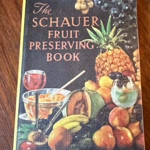 以下が含まれることがあります： 「ザ・シャウアー・フルーツ・プレザービング・ブック」というタイトルのヴィンテージの料理本。パイナップル、ブドウ、桃、メロンなど、さまざまな果物のカラフルなイラストが描かれている。