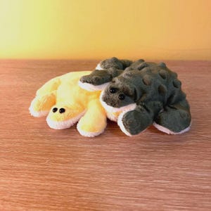 May include: Two plush toys: one yellow, the other dark green. The yellow toy has a rounded shape with black button eyes and white trim. The dark green toy has a bumpy texture and also features black button eyes and white trim. Both are on a wooden surface.