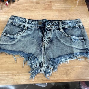Puede incluir: Pantalones cortos de mezclilla azul claro con bordes deshilachados y detalles desgastados. Los pantalones cortos tienen una cremallera y un bot&oacute;n.