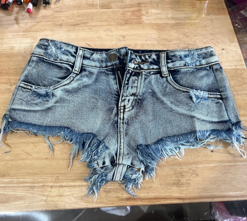 Puede incluir: Pantalones cortos de mezclilla azul claro con bordes deshilachados y detalles desgastados. Los pantalones cortos tienen una cremallera y un bot&oacute;n.
