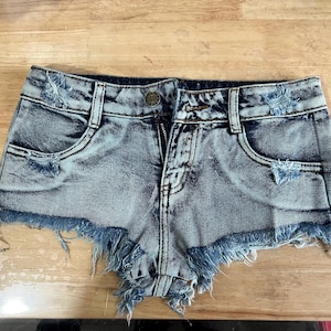 Puede incluir: Pantalones cortos de mezclilla azul claro con bordes deshilachados y detalles desgastados. Los pantalones cortos tienen una cremallera y trabillas para el cintur&oacute;n.