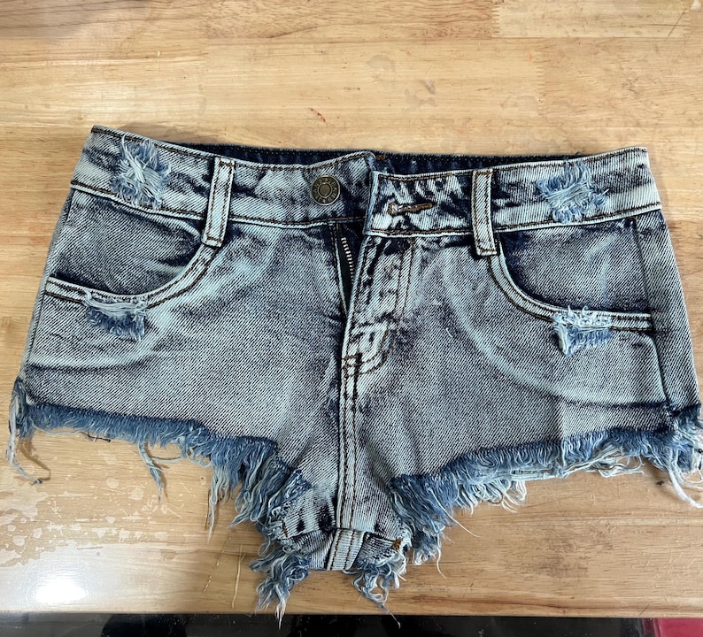Puede incluir: Pantalones cortos de mezclilla azul claro con bordes deshilachados y detalles desgastados. Los pantalones cortos tienen una cremallera y trabillas para el cintur&oacute;n.