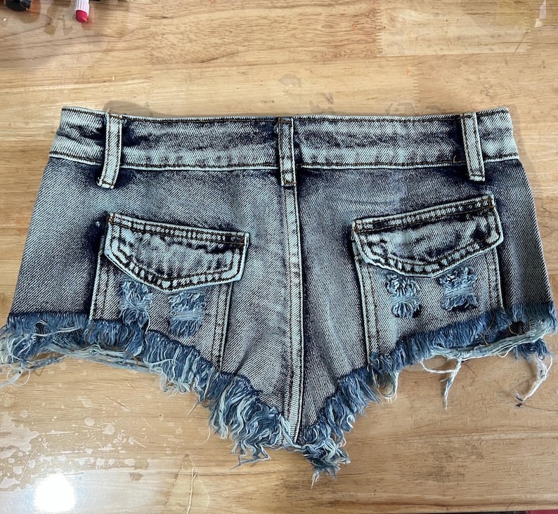 Puede incluir: Un par de pantalones cortos de mezclilla con dobladillo deshilachado y detalles desgastados. Los pantalones cortos tienen dos bolsillos en la parte posterior y un cierre de bot&oacute;n y cremallera en la parte delantera.