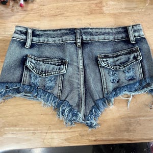 Puede incluir: Pantalones cortos de mezclilla azul claro con bordes deshilachados y detalles desgastados. Los pantalones cortos tienen dos bolsillos traseros y una cremallera.
