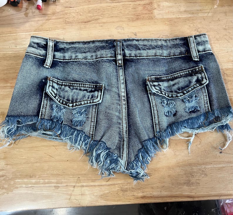 Puede incluir: Pantalones cortos de mezclilla azul claro con bordes deshilachados y detalles desgastados. Los pantalones cortos tienen dos bolsillos traseros y una cremallera.