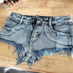 Puede incluir: Pantalones cortos de mezclilla azul claro con bordes deshilachados y detalles desgastados. Los pantalones cortos tienen cierre de bot&oacute;n y cremallera y dos bolsillos delanteros.