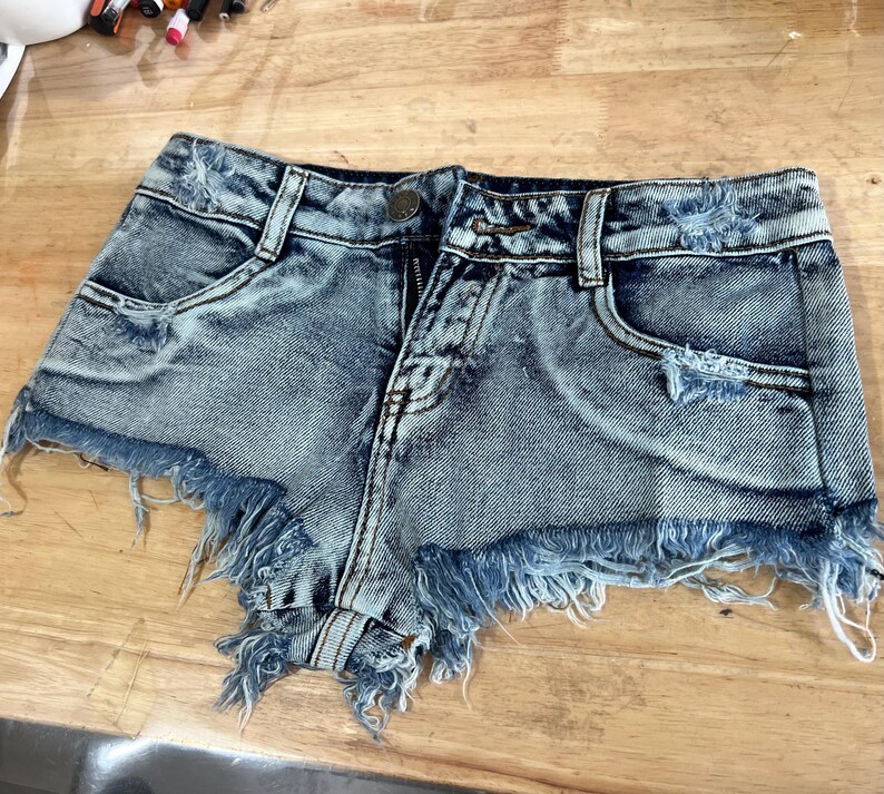 Puede incluir: Pantalones cortos de mezclilla azul claro con bordes deshilachados y detalles desgastados. Los pantalones cortos tienen cierre de bot&oacute;n y cremallera y dos bolsillos delanteros.