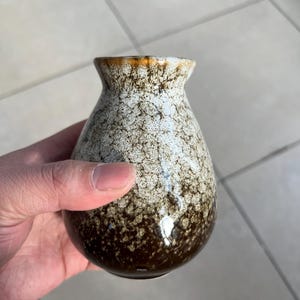 Op de afbeelding: Een kleine, bruin-witte keramische vaas met een gespikkeld glazuur. De vaas heeft een rond lichaam en een smalle hals.