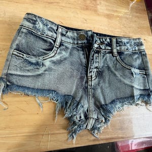 Puede incluir: Pantalones cortos de mezclilla azul claro con bordes deshilachados y detalles desgastados. Los pantalones cortos tienen una cremallera y un bot&oacute;n.