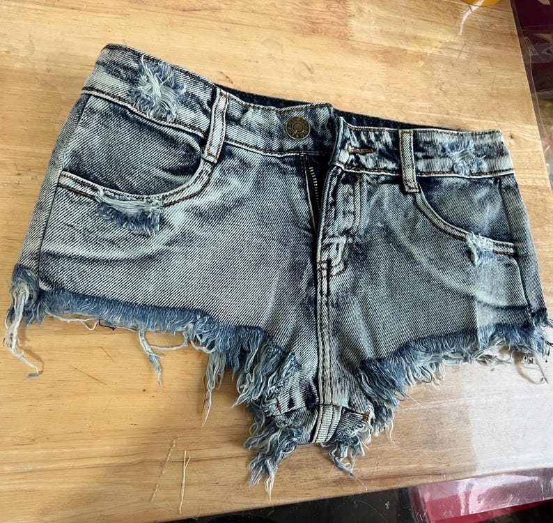 Puede incluir: Pantalones cortos de mezclilla azul claro con bordes deshilachados y detalles desgastados. Los pantalones cortos tienen una cremallera y un bot&oacute;n.