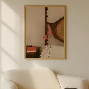 May include: A framed image featuring a bedroom scene. A wooden bed frame with a woven backboard is visible. A pink gingham bikini top hangs from the bedpost. A glass bottle and a red book sit on a wooden nightstand. A white pillow with a scalloped edge is on the bed.
